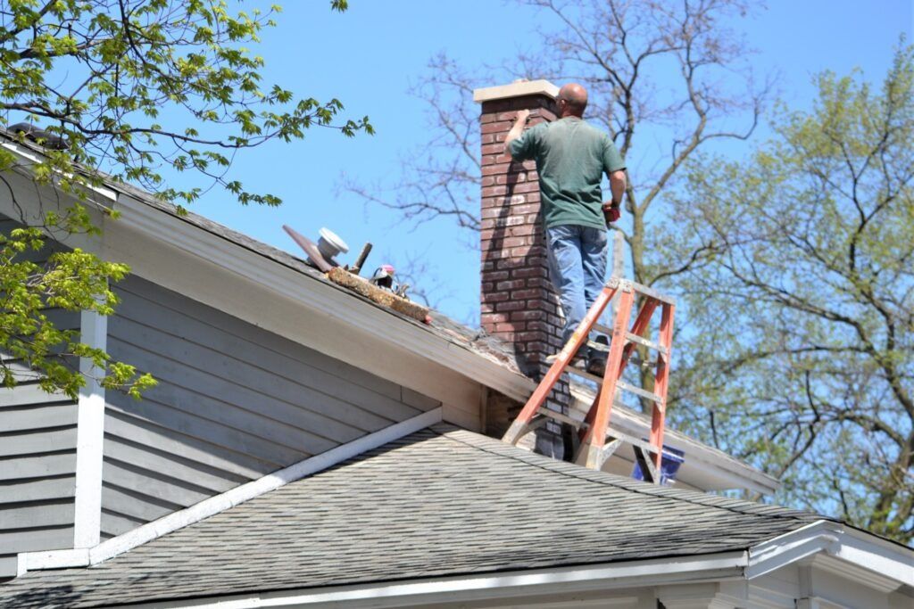 A person on a ladder repairs a brick chimney on a house roof. Blue sky, green trees.