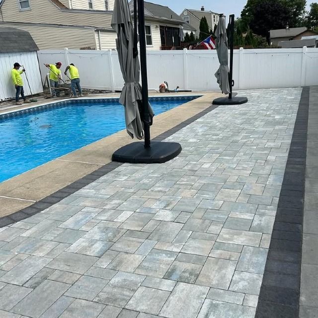 Workers near a pool with a paved patio. The patio is gray with a dark border; two umbrellas are standing upright.
