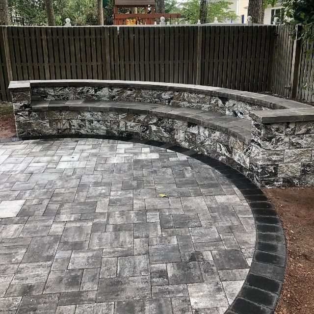Curved stone seating built into a raised stone wall on a gray paver patio. A wooden fence is in the background.