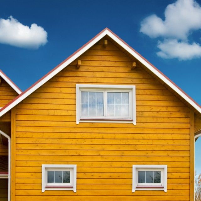 Yellow wooden house with a red roof against a blue sky with fluffy clouds. Two small windows below a larger one.