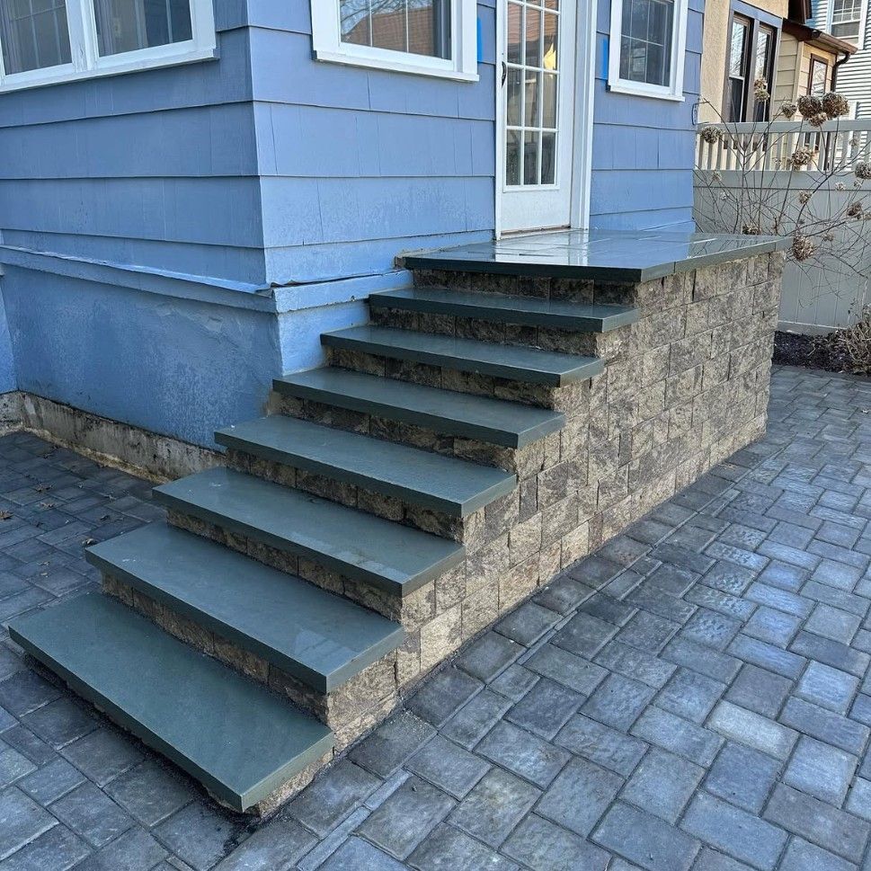Blue house with stone steps leading to a white door. The steps are gray and lead from a paver patio.