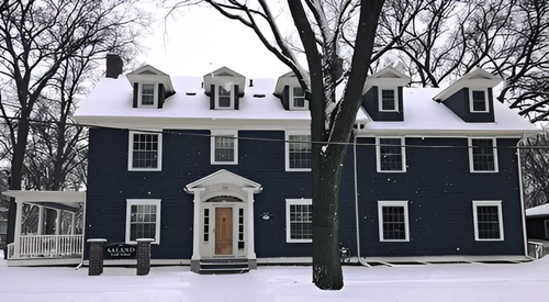 Snow-covered dark blue house with white trim and a large tree in front.