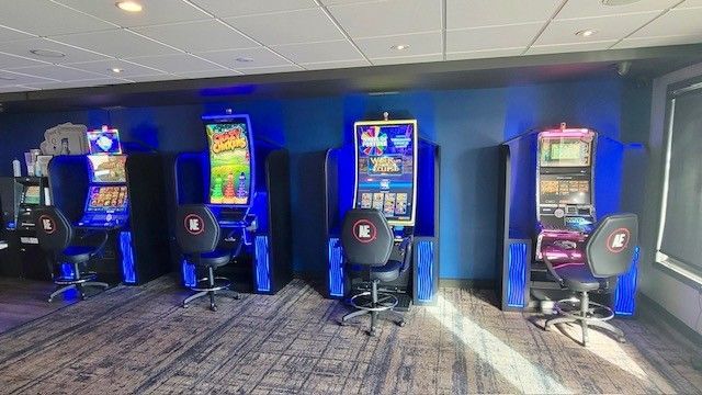 Slot machines in a row, each with a chair. Blue and pink lighting, in a casino setting.