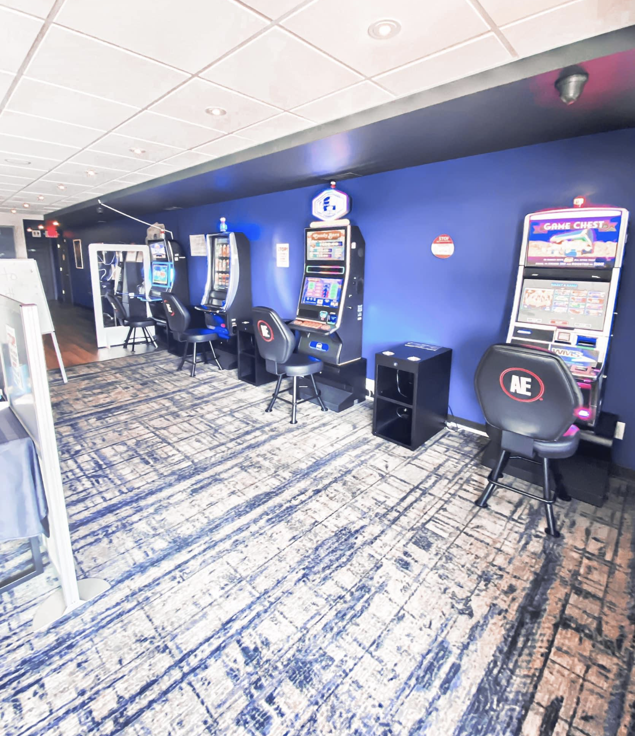 Gaming area with slot machines, blue walls, patterned carpet, and black chairs.