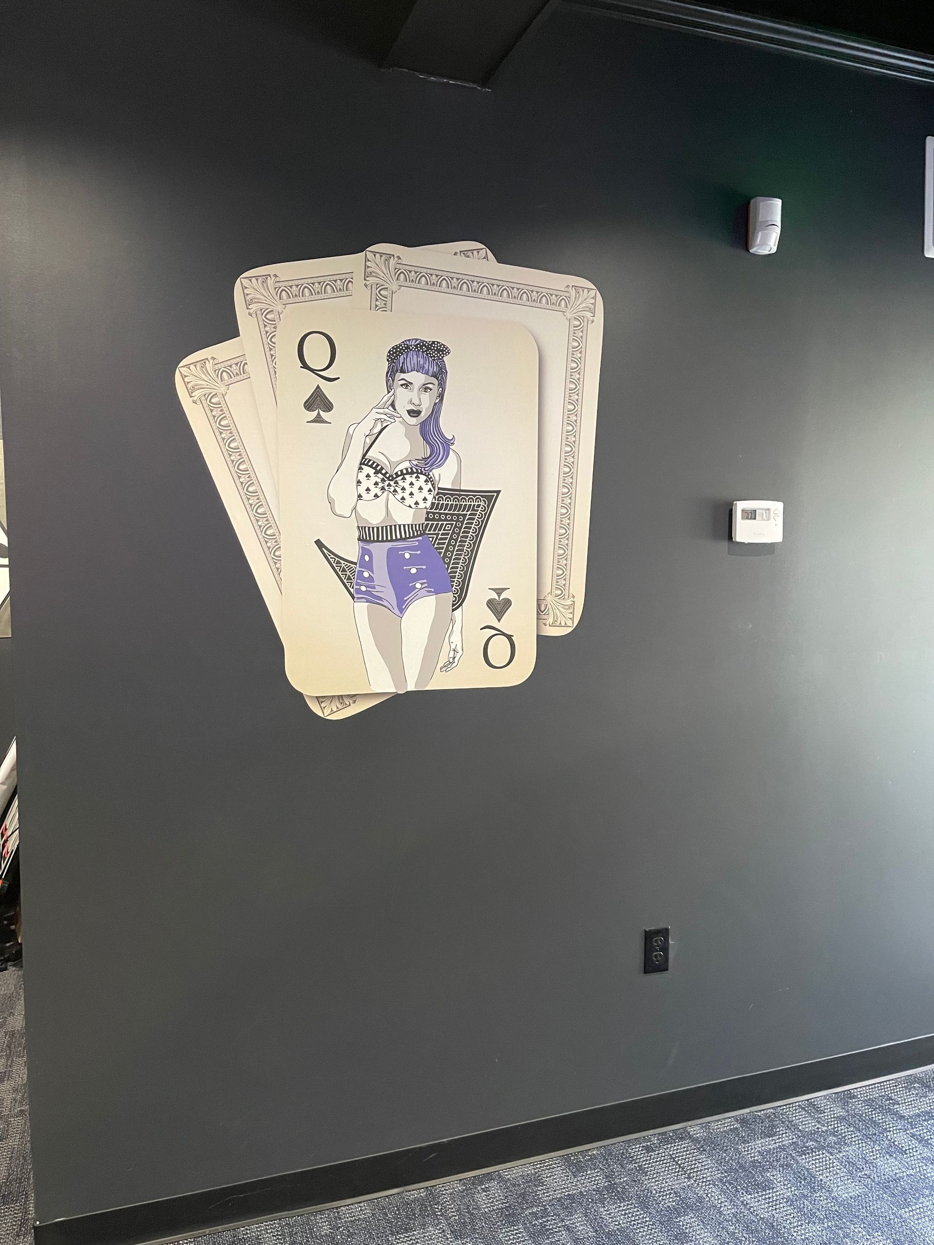 A wall art piece depicting the Queen of Spades card with illustration, displayed on a dark gray wall.