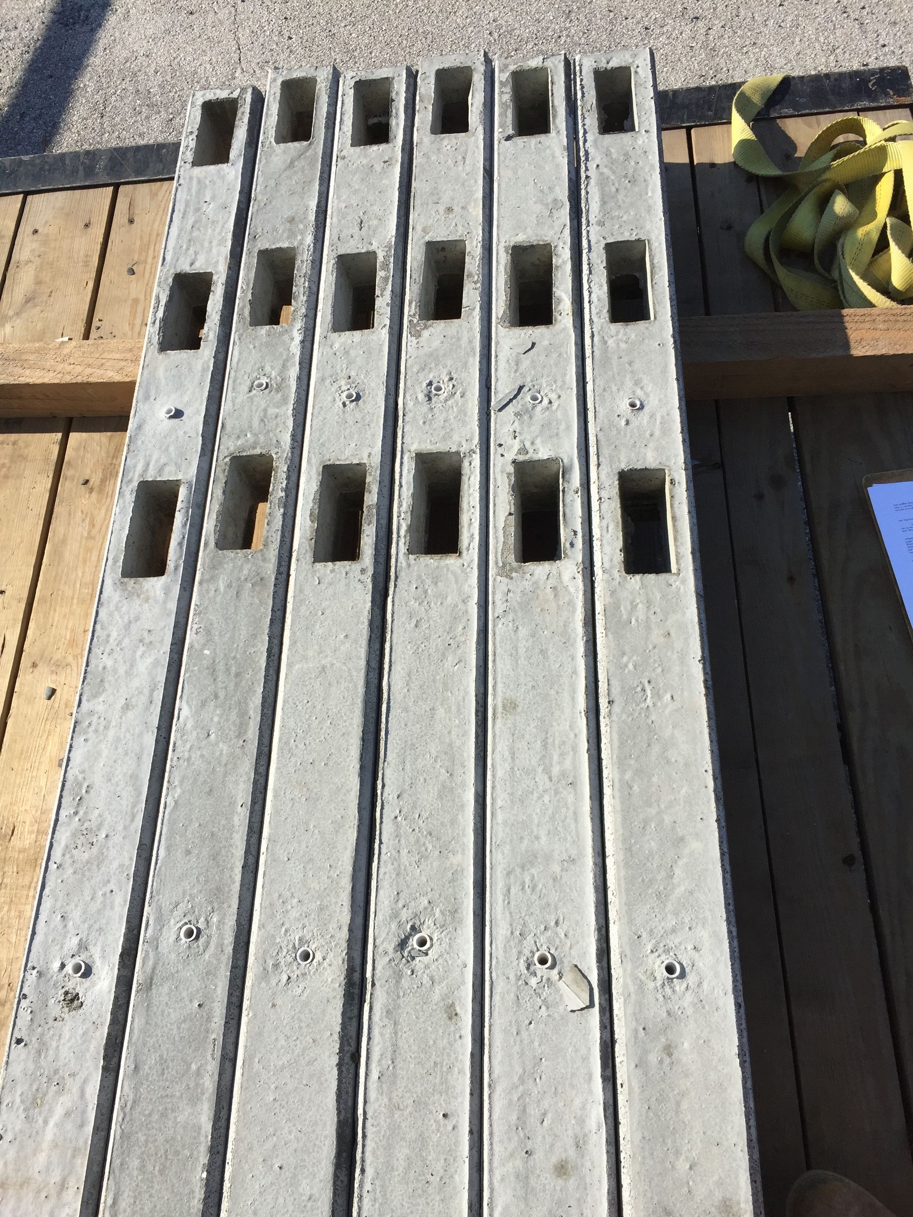 Six long, grey concrete posts with rectangular cutouts, resting on a wooden trailer bed outdoors.