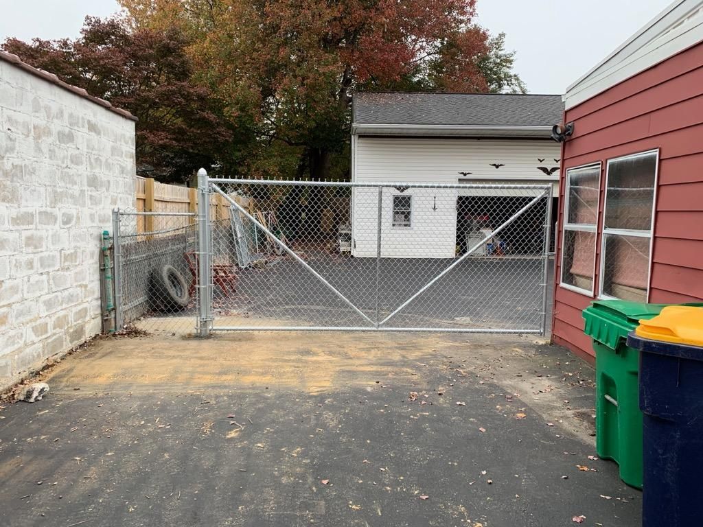 A metal chain-link gate spans a driveway between a light-colored brick wall and a red building, with trees in the back.