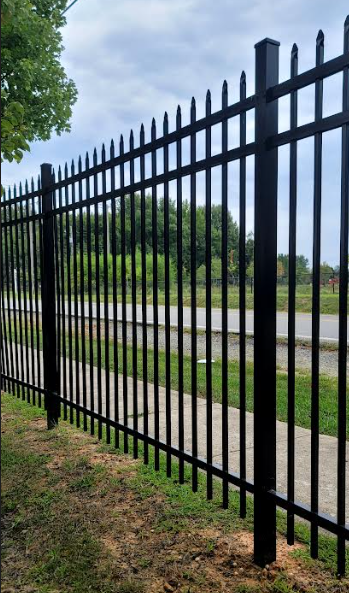 A black metal picket fence with spear-point finials borders a sidewalk and grassy area under a cloudy sky.