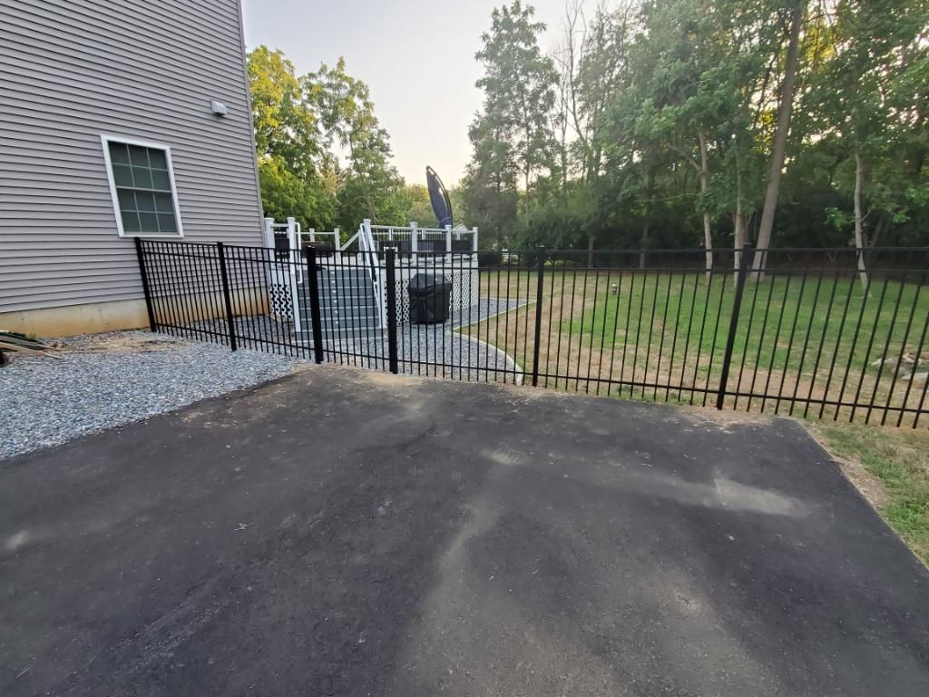 A black metal fence separates a paved driveway and gravel area from a grassy backyard with a deck.