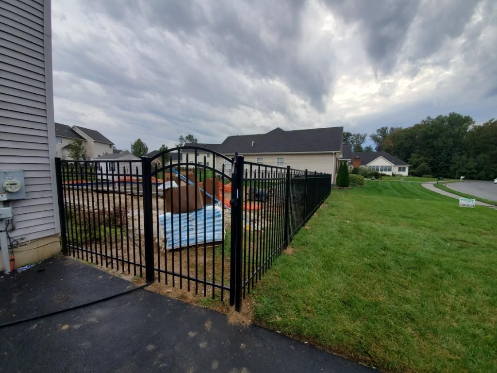 A black metal fence with a gate encloses a backyard with construction materials on a lawn beside a suburban home.