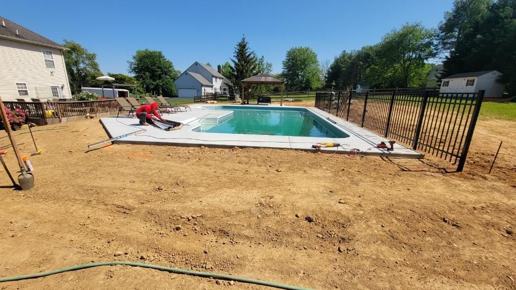 A person in a red shirt works on the stone pool deck of a backyard swimming pool surrounded by dirt and a metal fence.