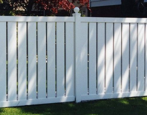 A white vinyl privacy fence with vertical pickets and a decorative ball-topped post, standing on a green grass lawn.