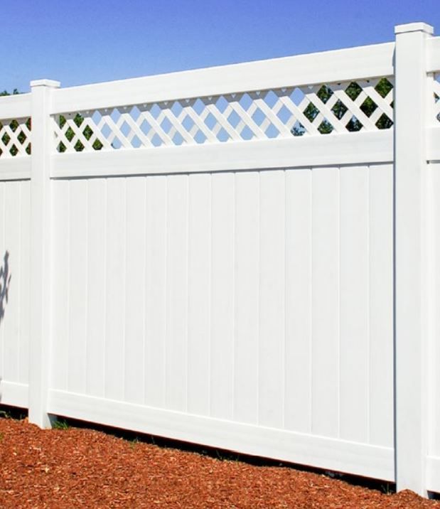 A white vinyl privacy fence with a lattice top design standing over a patch of brown mulch against a blue sky.