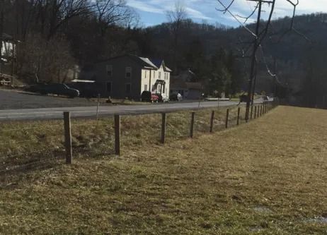 A wooden fence line running along a grassy field next to a road, with a two-story house in the background.