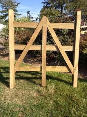 A wooden gate with a triangular frame design stands between two posts in a grassy area with shrubs in the background.