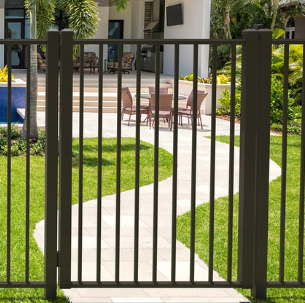 A dark bronze vertical-bar metal fence gate stands in front of a stone walkway leading to a patio with outdoor furniture.