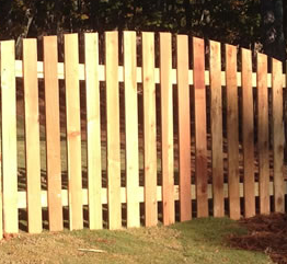 A wooden picket fence with a gently arched top design, set against a background of grass and trees.