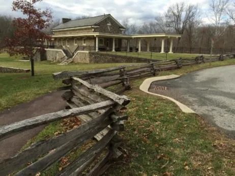 A split-rail wooden fence lines a paved path leading toward a stone visitor center building with a colonnaded porch.