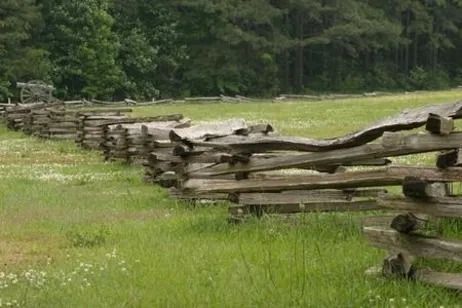 A rustic wooden split-rail fence stretches across a green grassy field toward a line of trees.