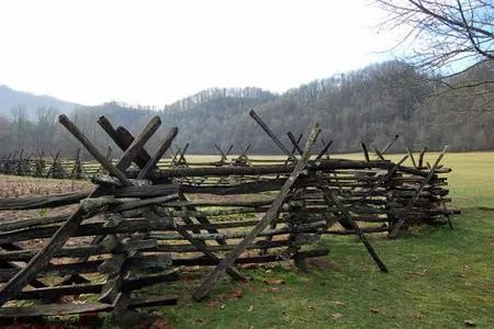 A rustic zigzag wooden fence stretches across a green field toward tree-covered mountains under a bright, overcast sky.