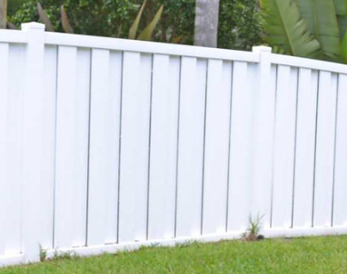 A white vinyl privacy fence stands in a backyard against a backdrop of green foliage and grass.