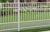 A white, vertical-bar vinyl fence section with a dual-spaced picket design, set against a green lawn and house.