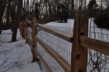 A split-rail wooden fence with wire mesh lines a snowy yard containing a small playset at dusk.