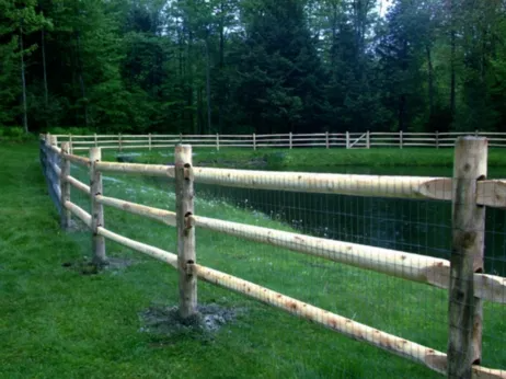 A rustic, three-rail wooden post-and-rail fence with wire mesh backing curves along the edge of a pond near a forest.