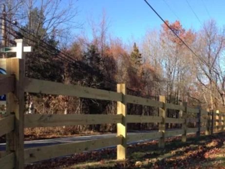 A wooden split-rail fence runs along a paved path bordered by trees under a clear blue sky.