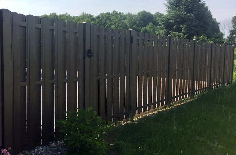 A brown, vertical-slat fence with a gate, installed outdoors in a grassy area with trees in the background.