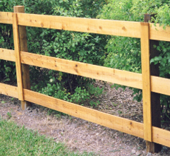 A wooden, three-rail fence stands in front of green bushes, with a patch of grass in the foreground.