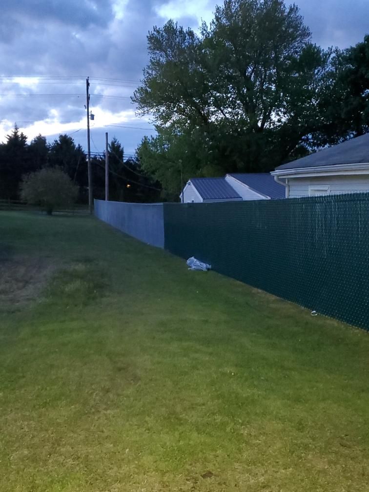A long, green privacy fence runs along a grassy lawn next to white buildings under a cloudy, late afternoon sky.