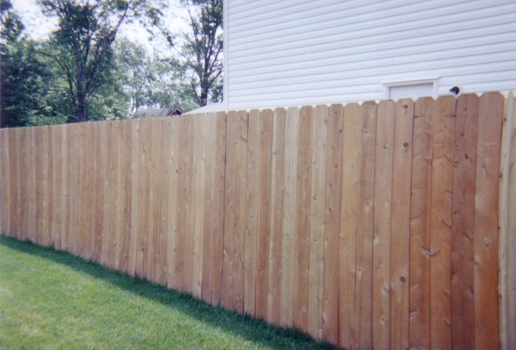 A tall, light-brown wood privacy fence spans across a green lawn in front of a white house.
