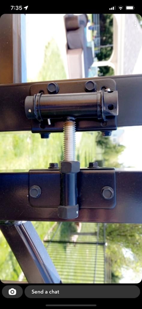 Black adjustable gate hinge mounted on horizontal metal rails, featuring a central tension bolt and mounting plates.