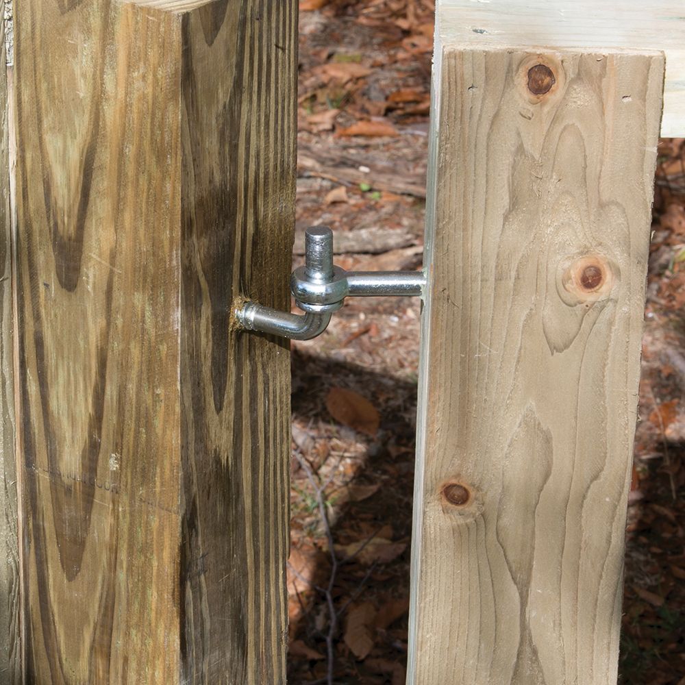 A metal bolt-hook hinge connects a wooden gate post to a wooden gate, seen from a close-up angle.