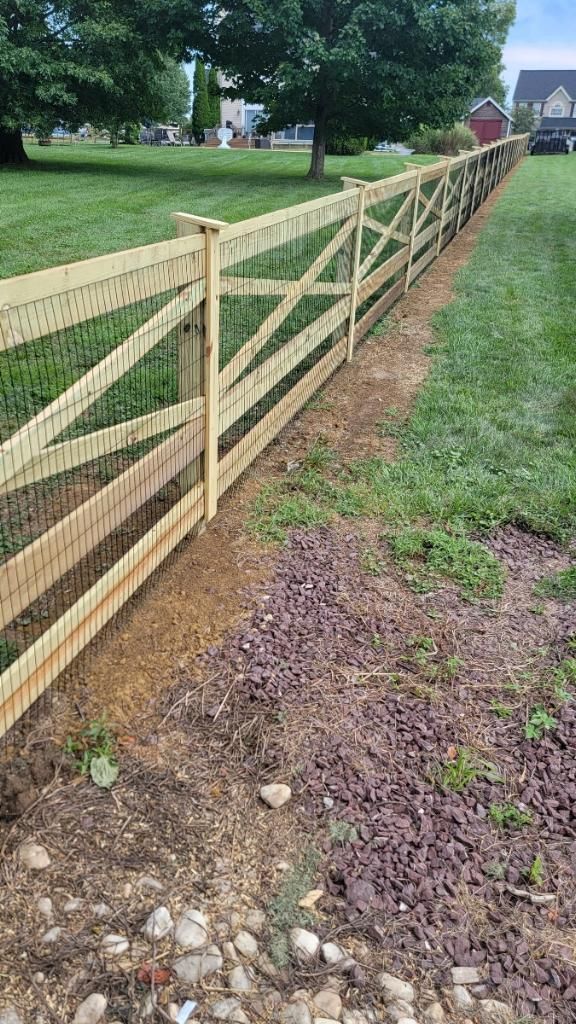 A new wooden rail fence with attached wire mesh runs along a grassy yard bordering a garden bed with rocks and mulch.