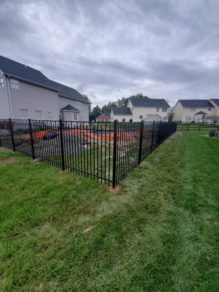 A black metal fence encloses a backyard area featuring orange construction netting and a dirt surface under a cloudy sky.