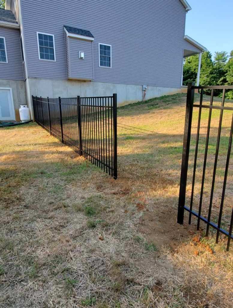 A black metal fence runs across a dry, grassy yard beside the side wall of a gray, two-story house.