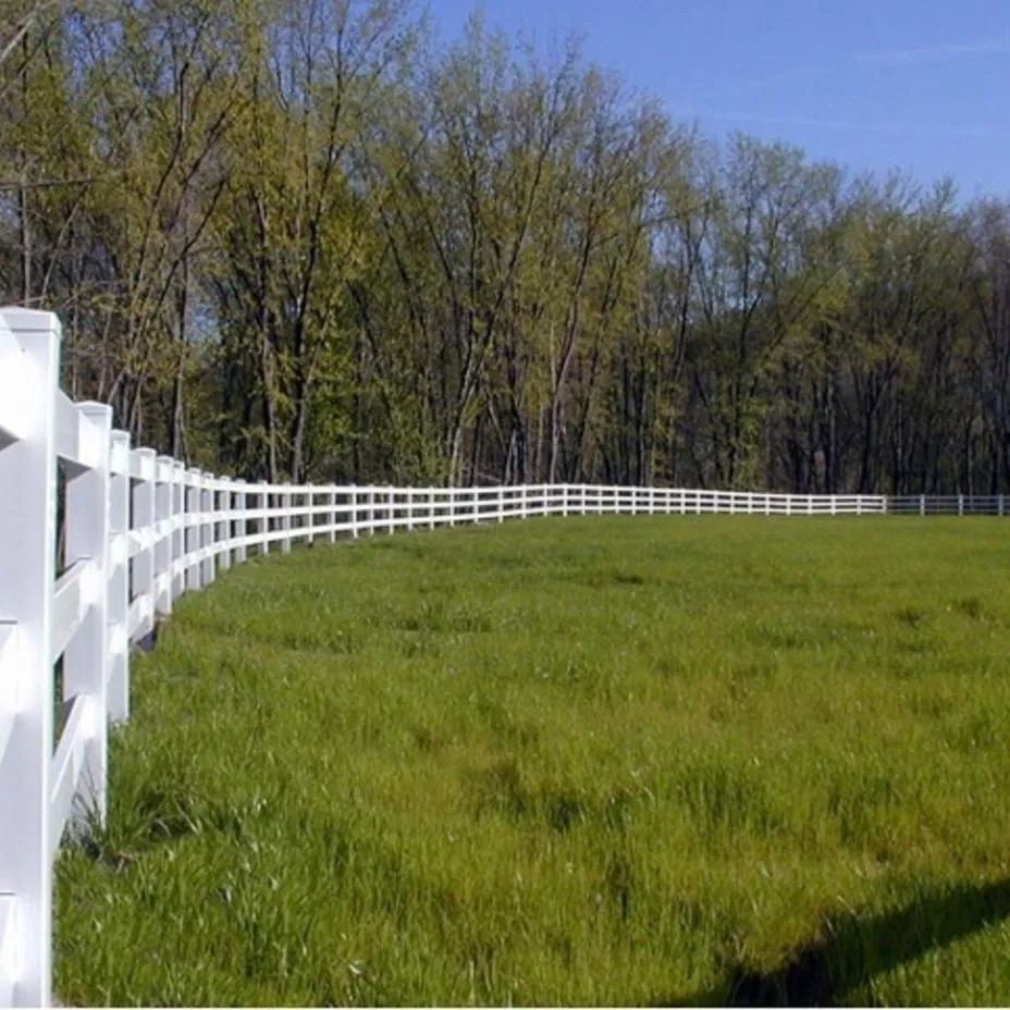 A curved white fence borders a lush green field in front of a forest of trees under a clear blue sky.