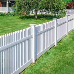 A white vinyl picket fence stretches across a green lawn in front of a tree and a house.