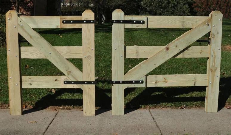 Two wooden farm-style gates standing side-by-side with black metal hinges, set against a green lawn and concrete path.