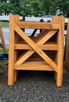 A stack of unfinished wooden gate frames with diagonal cross-bracing, resting on a gravel surface outdoors.
