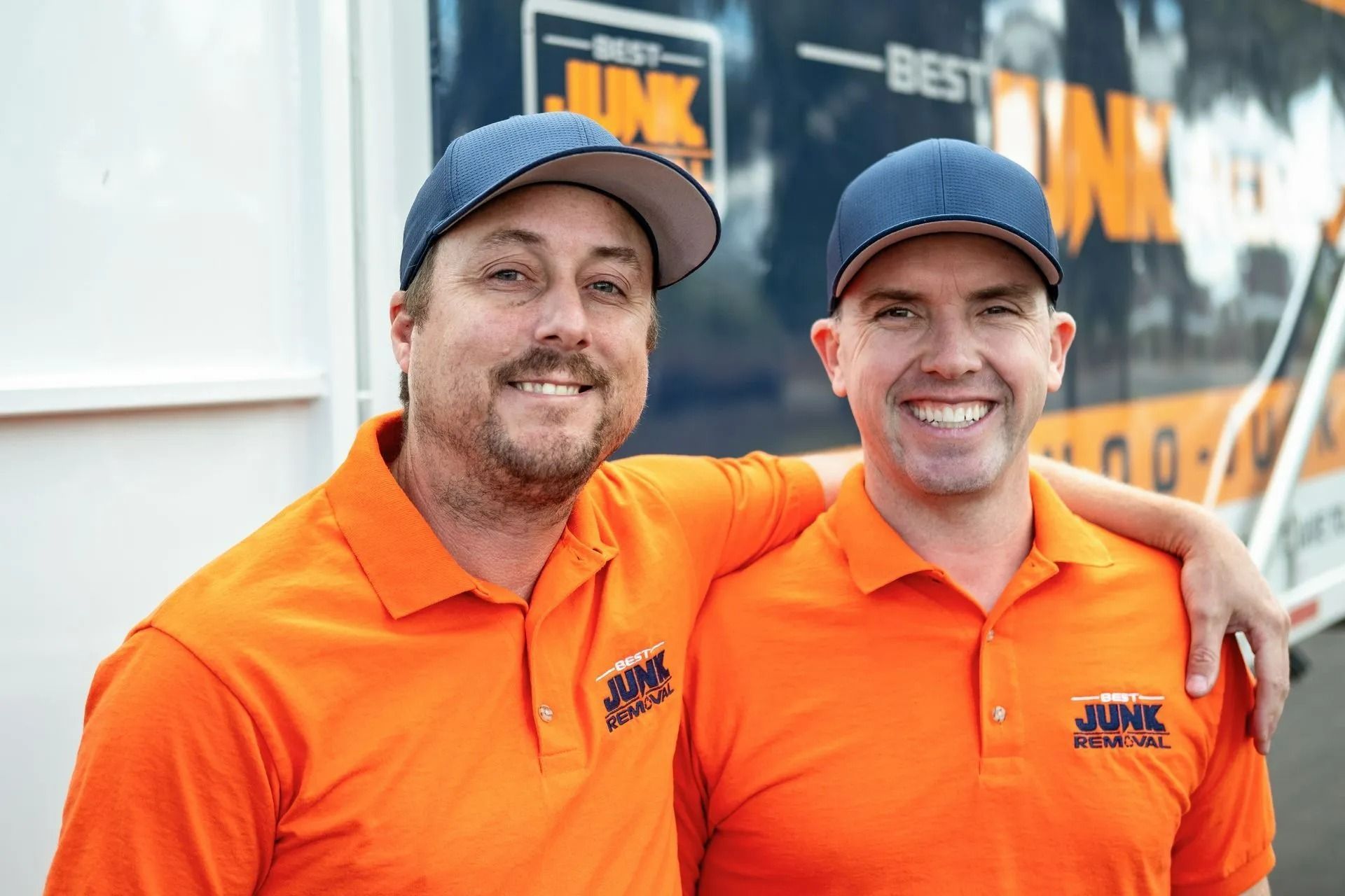 Two men smiling, wearing orange shirts and blue caps, with 