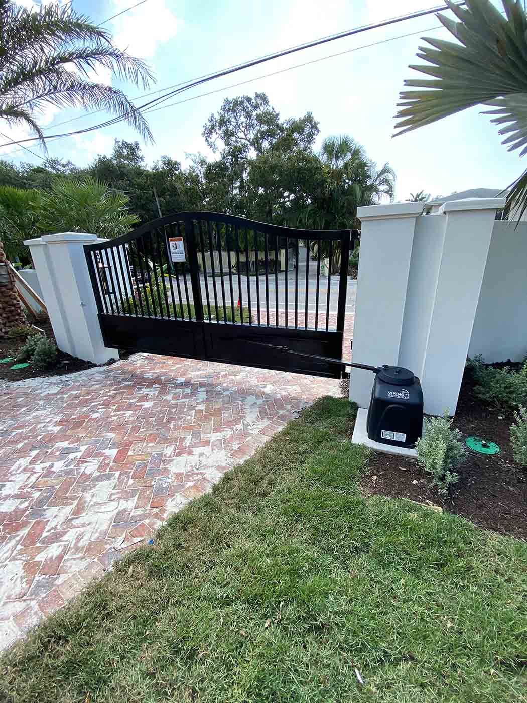 A black gate is sitting on top of a brick driveway next to a palm tree.