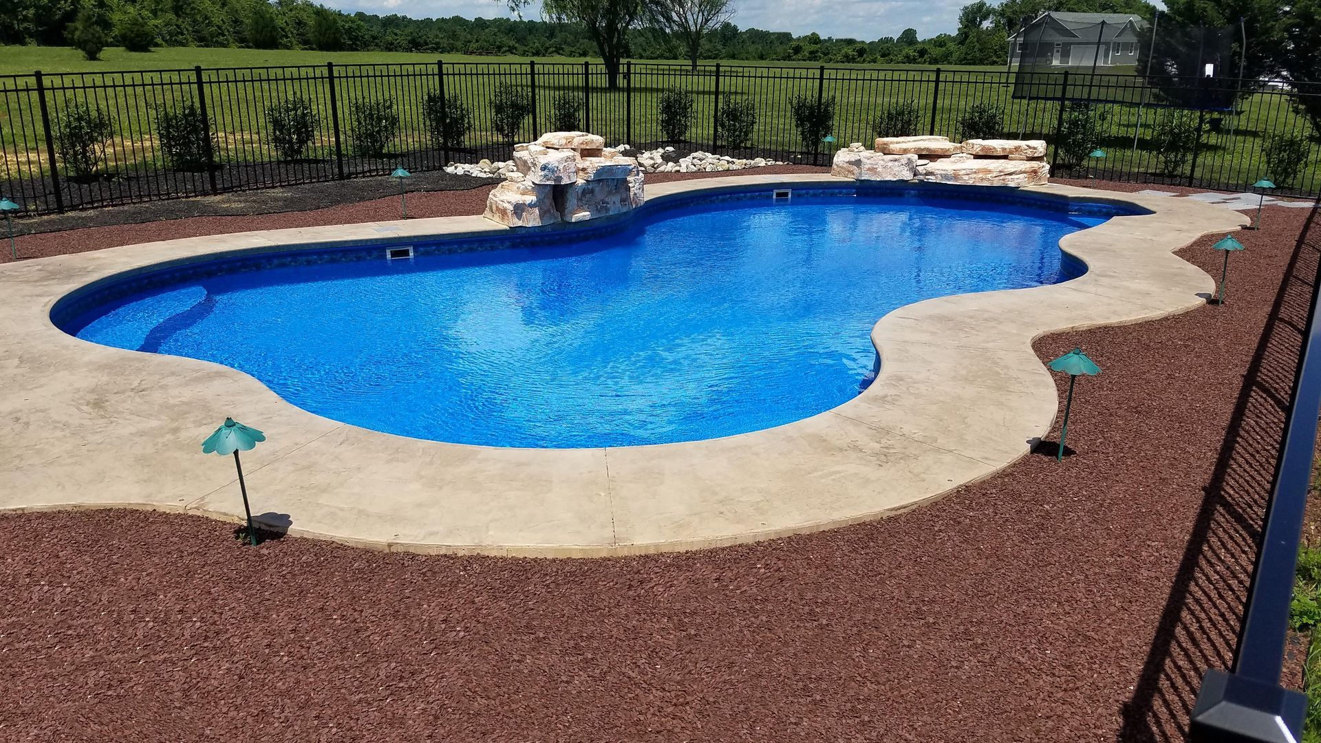 A large blue swimming pool is surrounded by gravel and a fence.