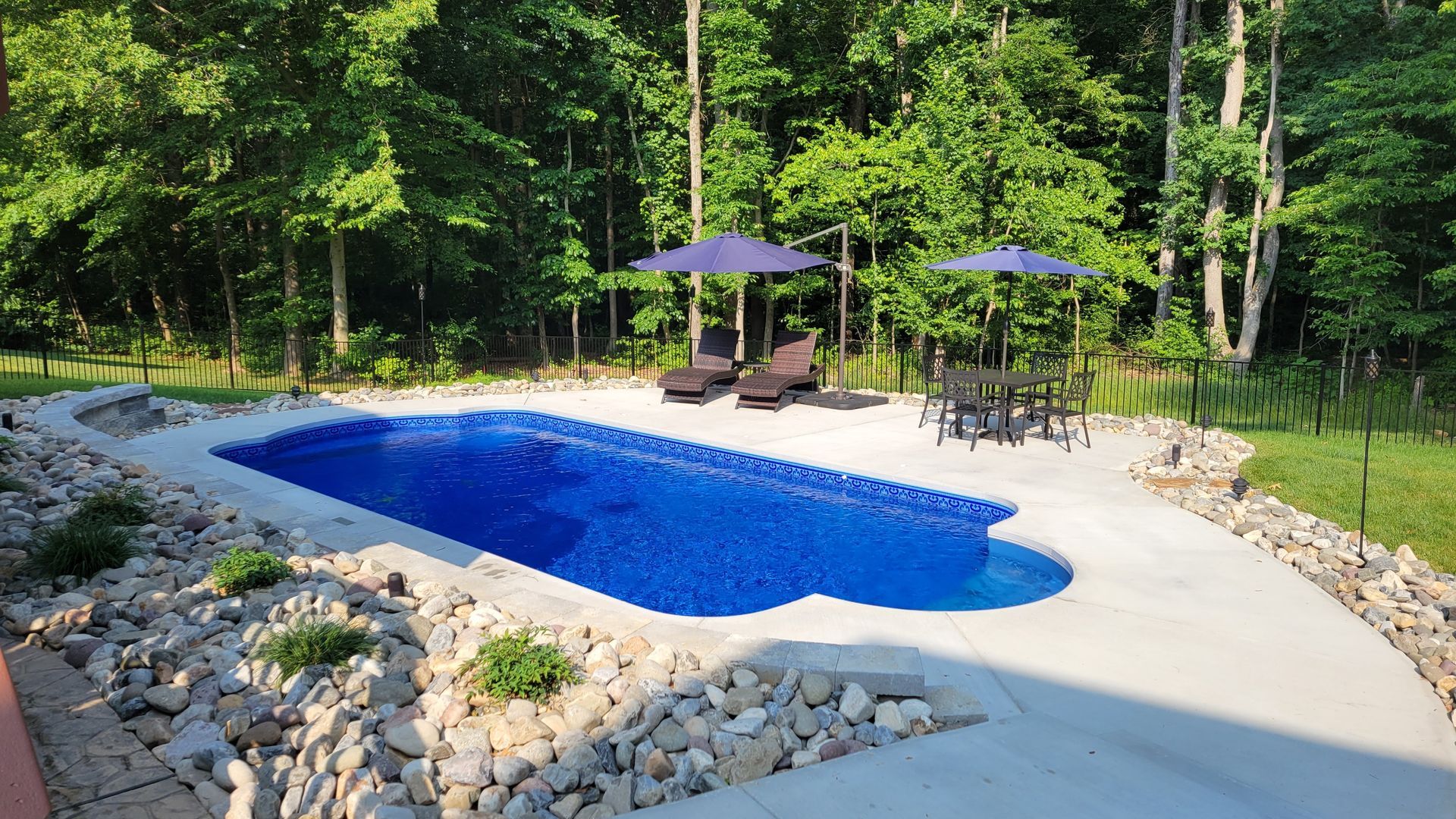 A large blue swimming pool with umbrellas and chairs in a backyard surrounded by trees.