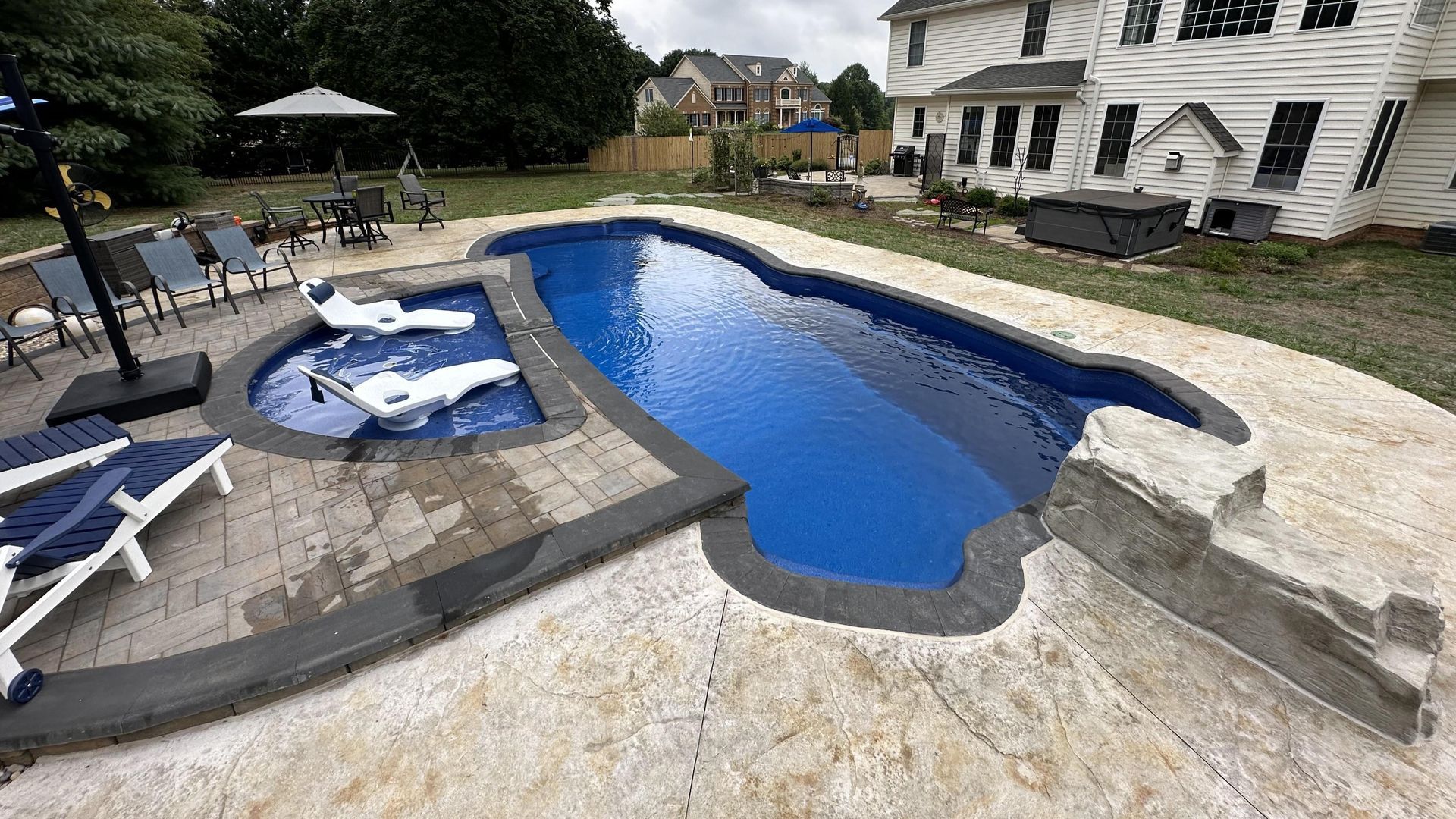 Backyard pool with blue water, stone deck, lounge chairs, and a hot tub beside a white house
