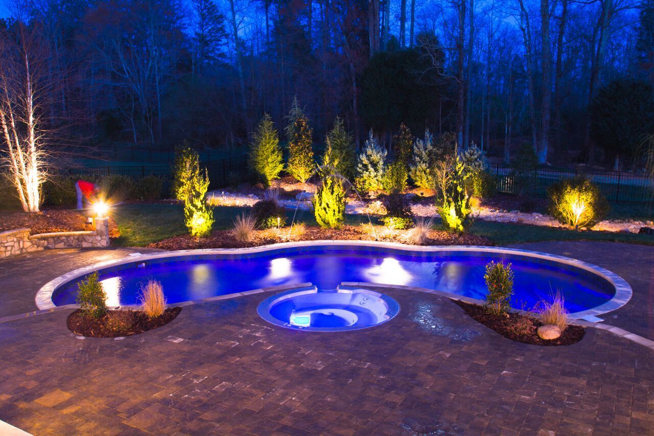 Night view of a swimming pool with blue lighting surrounded by landscape lighting and trees.