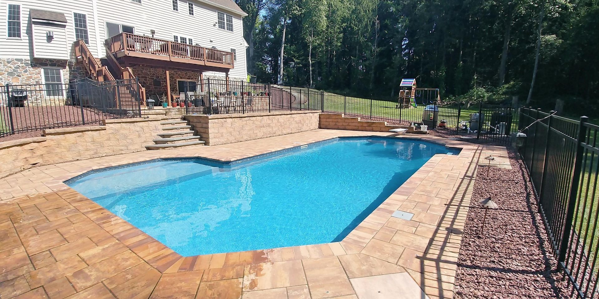 Backyard with a pool, brick patio, and a two-story house with a wooden deck. Black fence surrounds the pool and yard.
