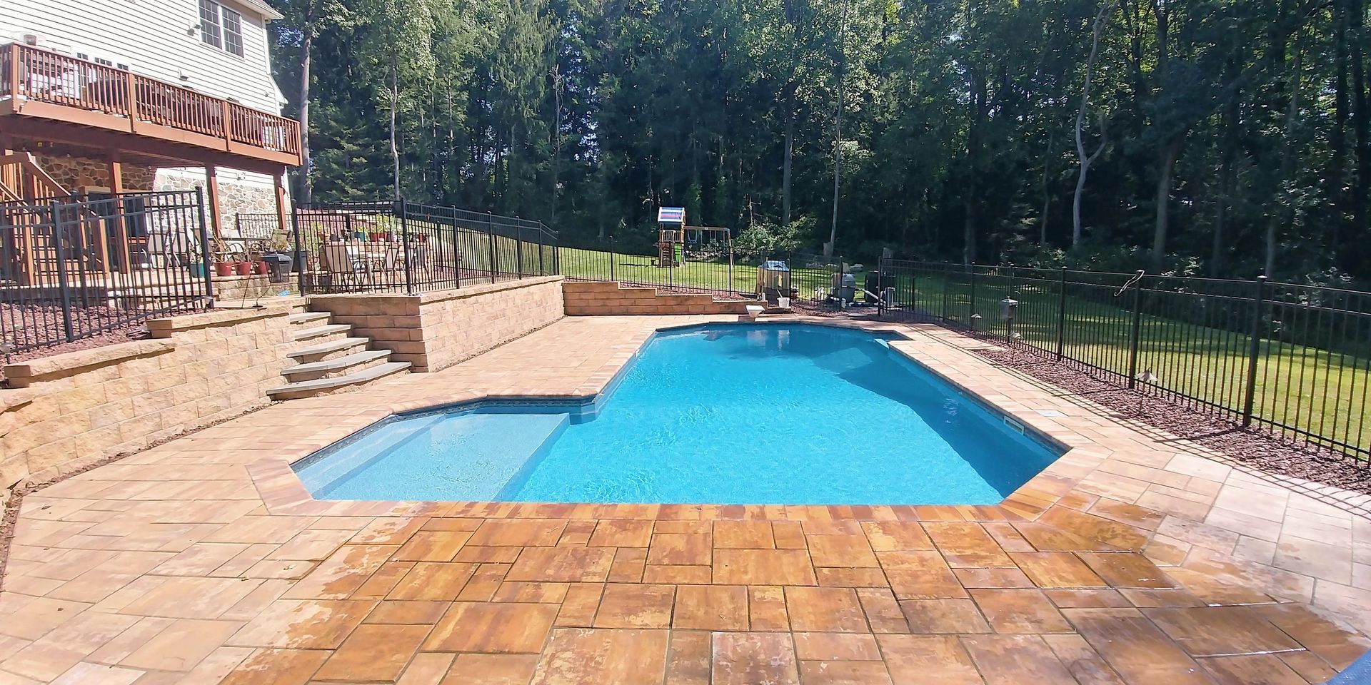 Swimming pool surrounded by brick patio, black fence, and trees. A large house with a wooden deck is in the background.
